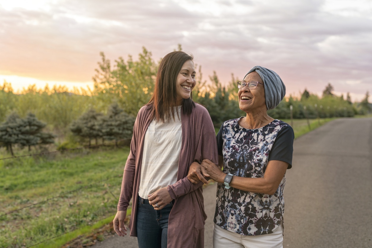 A senior woman with cancer walks with her adult daughter at sunset down a rural road. They are relaxing and staying active together. The affectionate pair are talking and walking with arms linked.